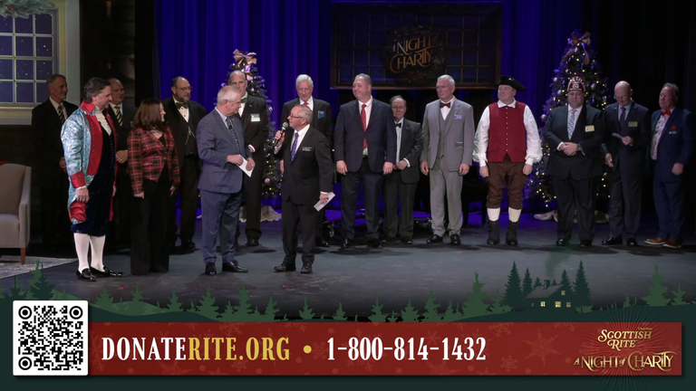 A group of people stand on a decorated stage during a holiday-themed charity event,  some individuals are speaking into microphones while others stand in a row behind them.