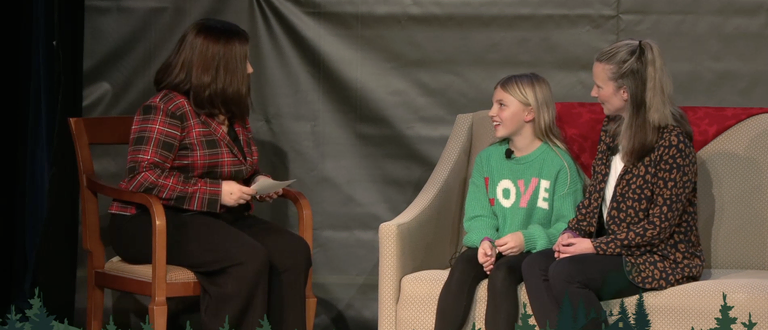 A woman in a plaid jacket sits in a chair interviewing a young girl and an adult woman seated together on a couch on stage.