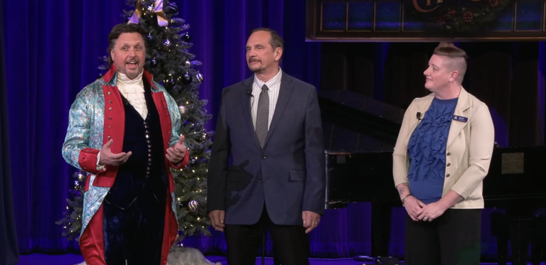 A man in a colorful costume and two presenters stand on a decorated stage with Christmas trees and a piano in the background.