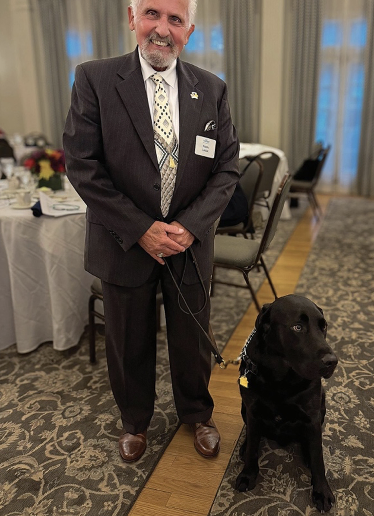 A smiling man in a suit and tie holding a Black Labrador dog by a leash
