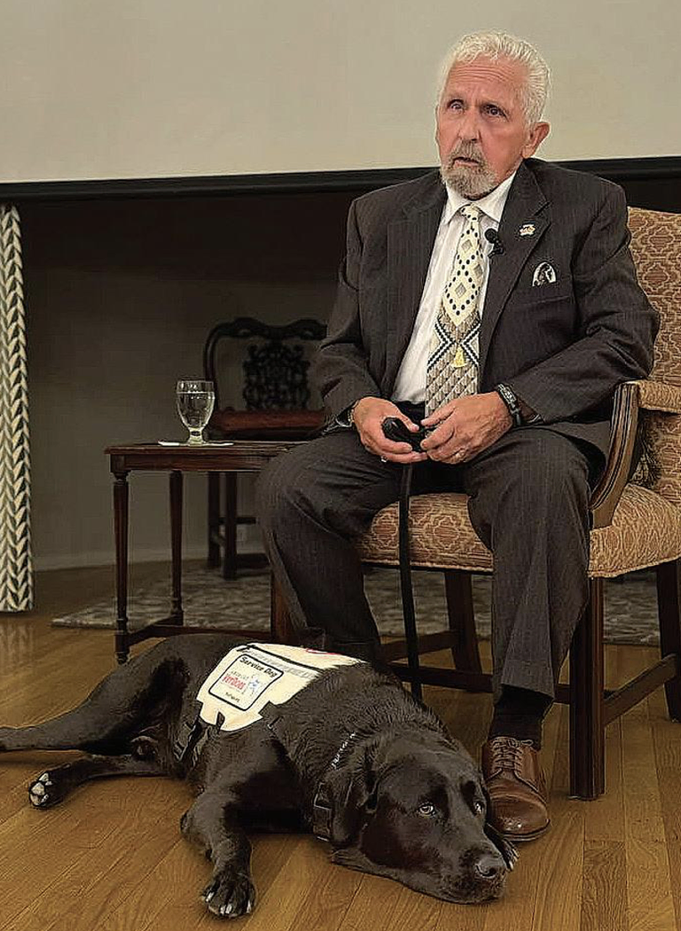 A man sitting down holding the leash of a Black Labrador dog, who is resting on the ground