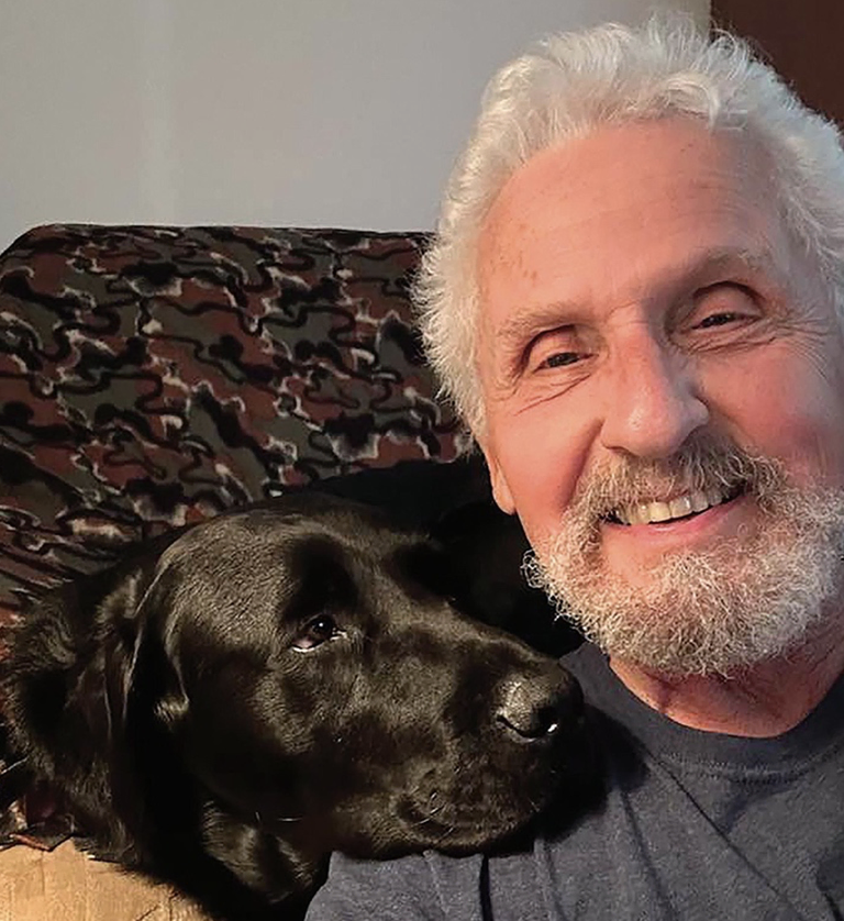 A selfie of a man smiling next to a Black Labrador dog, whose head is resting on the man’s shoulder
