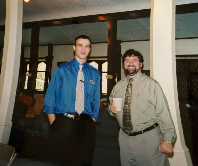 A younger man on the left, wearing a blue shirt and tie, while standing beside another man on the right, inside a Masonic lodge