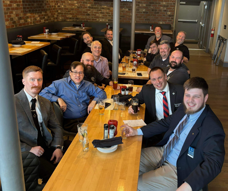 A large group of men sitting together at a long restaurant table, smiling and talking over drinks in a warm, brick-walled pub setting.
