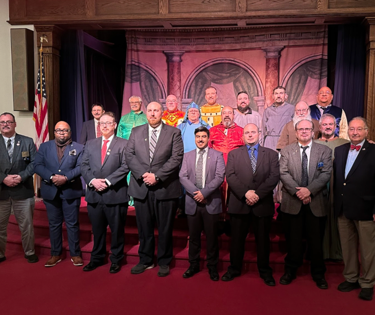 Two rows of Scottish Rite Brothers standing together in a lodge room; the front row is dressed in suits, while the back row wears colorful degree costumes.