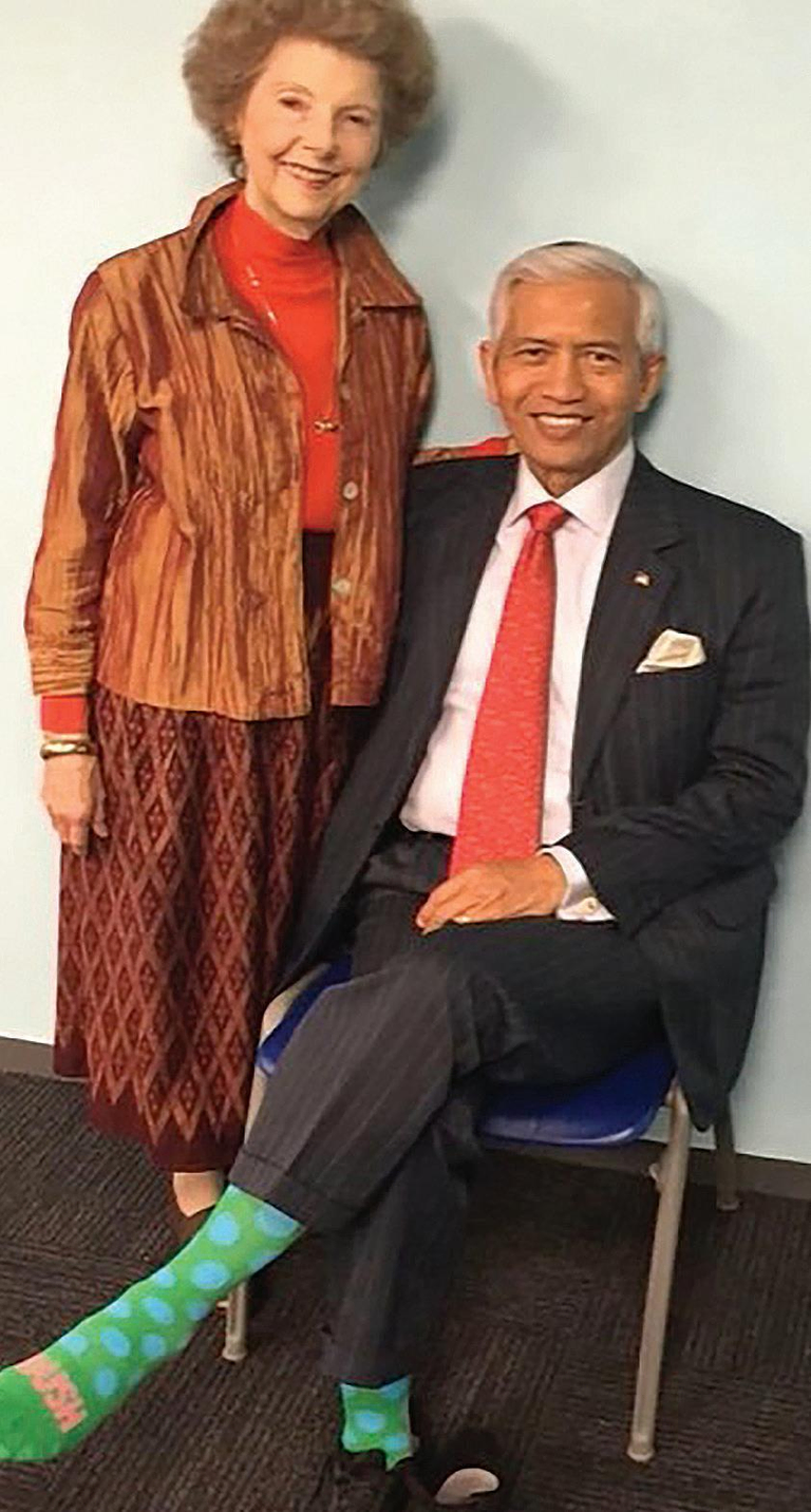 A woman dressed in a brown skirt stands next to a man sitting in a black suit with a red tie.