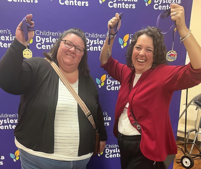 Two women, Michelle and Lindsey, stand in front of a Children’s Dyslexia Center backdrop, smiling and holding up medals in celebration.