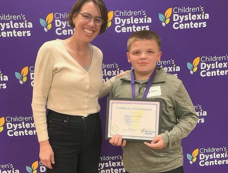A smiling tutor and a young boy named Gunner stand in front of a Children’s Dyslexia Center backdrop. Gunner is holding a framed certificate and wearing a medal around his neck.