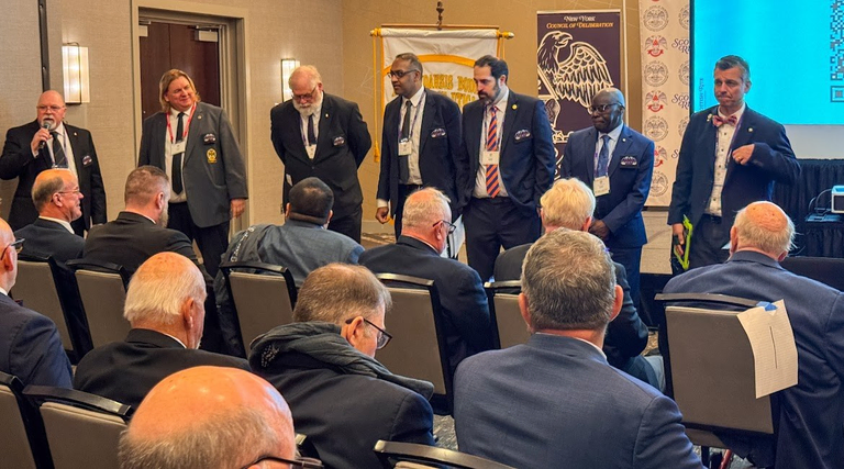 Seven men in suits stand at the front of a meeting room addressing attendees during a Scottish Rite session