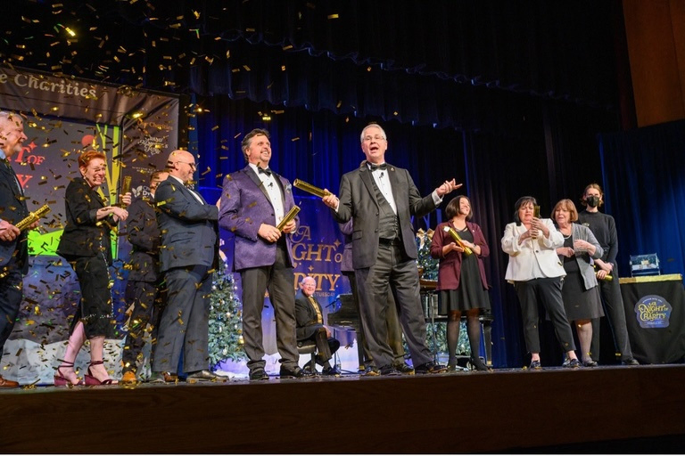 A group of men and women dressed in formal attire, clapping and celebrating on a stage surrounded by gold confetti