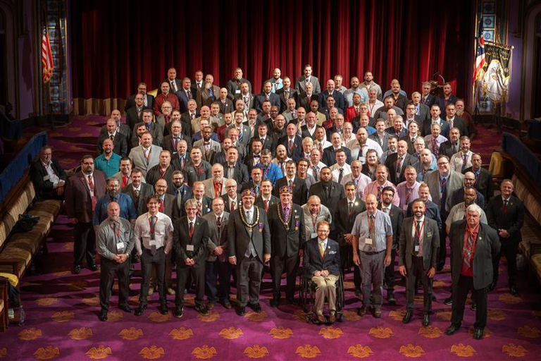 A large group of smiling Freemasons in business casual attire standing together in a lodge room in front of a stage adorned with a red curtain