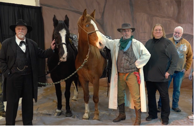 Alt text: Four men, two wearing Western attire, on a movie set with two horses