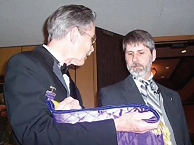 Two men standing next to each other, one presenting a Masonic apron to another.