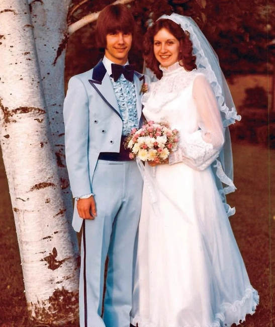 A young man in a formal blue suit standing next to a young woman in a wedding dress, holding a bouquet.